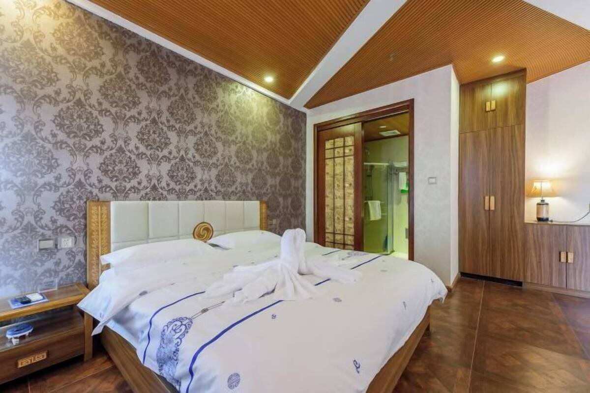 A hotel room with a bed, wooden headboard, patterned wallpaper, a wooden wardrobe, and a nightstand.
