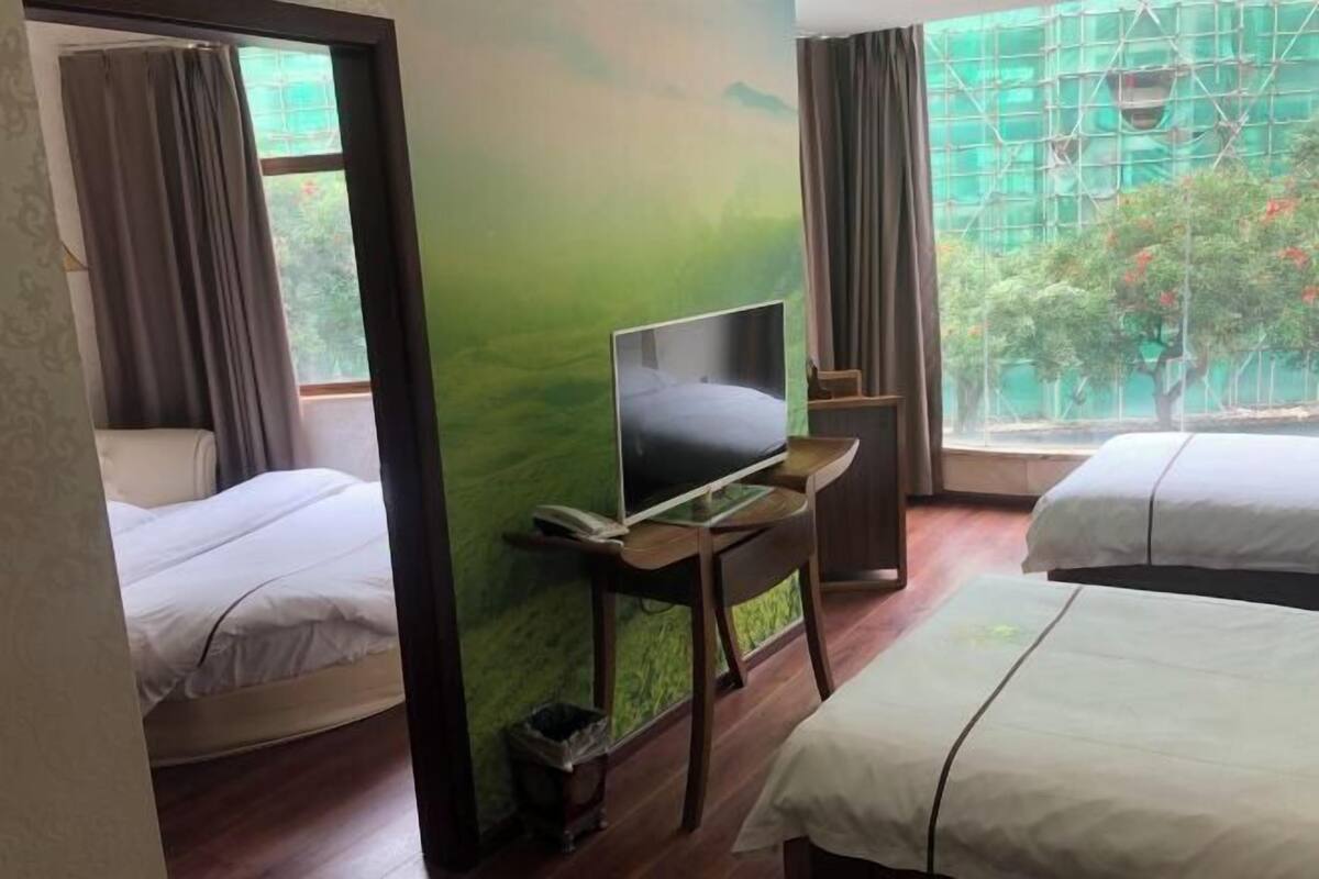 A hotel room with two beds, a TV on a stand, a window with a view of greenery, and a door leading to another room.