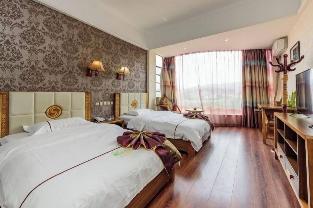 A hotel room with two beds, a wooden floor, a large window with curtains, and a television on a wooden stand.