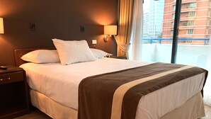A hotel room with a large bed, wooden nightstands, and a view of a building through the window. - Apart Hotel Elite  (Santiago)