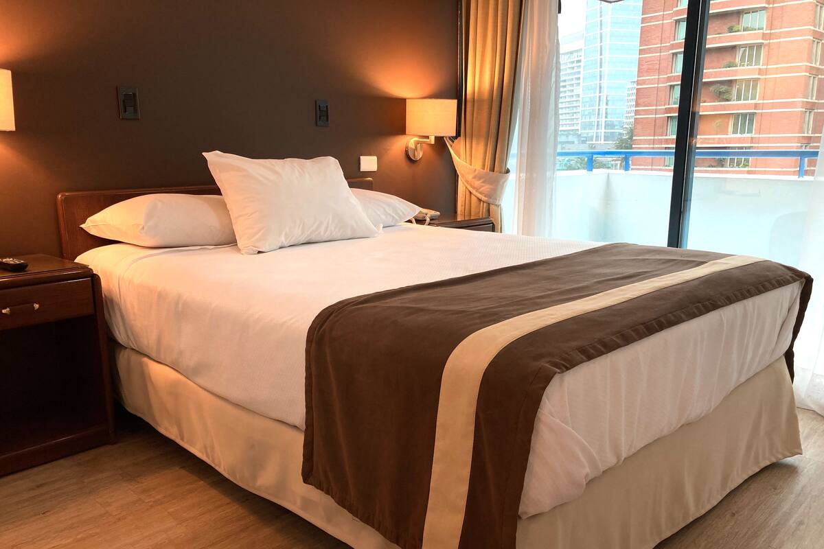 A hotel room with a large bed, wooden nightstands, and a view of a building through the window.