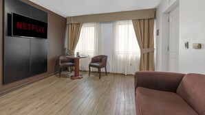A hotel room with a large flat-screen TV mounted on the wall, a wooden floor, a brown leather sofa, two chairs, a small round table, and a window with beige curtains. - Apart Hotel Elite  (Santiago)