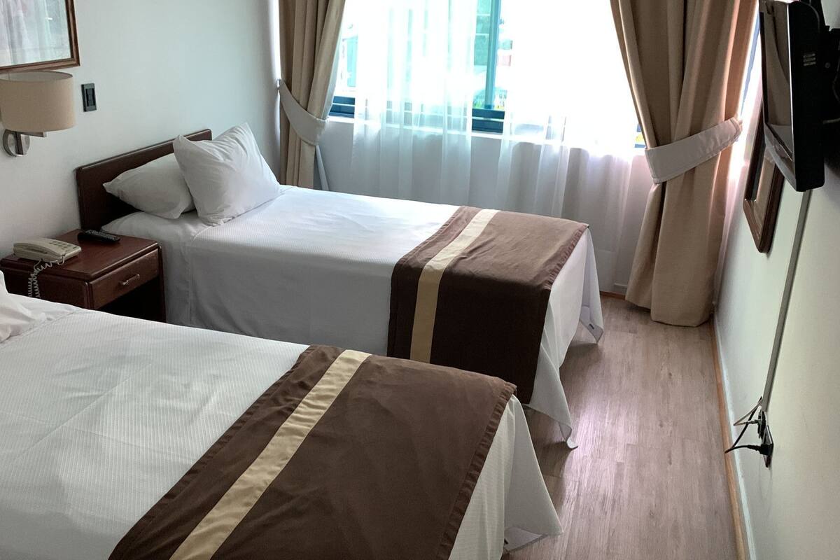 A hotel room with two beds, a bedside table with a lamp, and a window with curtains.