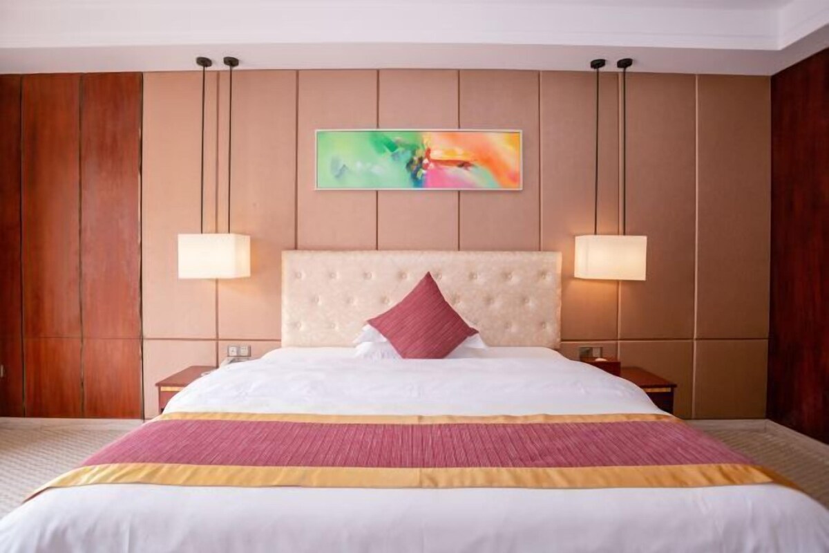 A hotel room with a large bed, two bedside lamps, a headboard, and a colorful abstract painting on the wall.