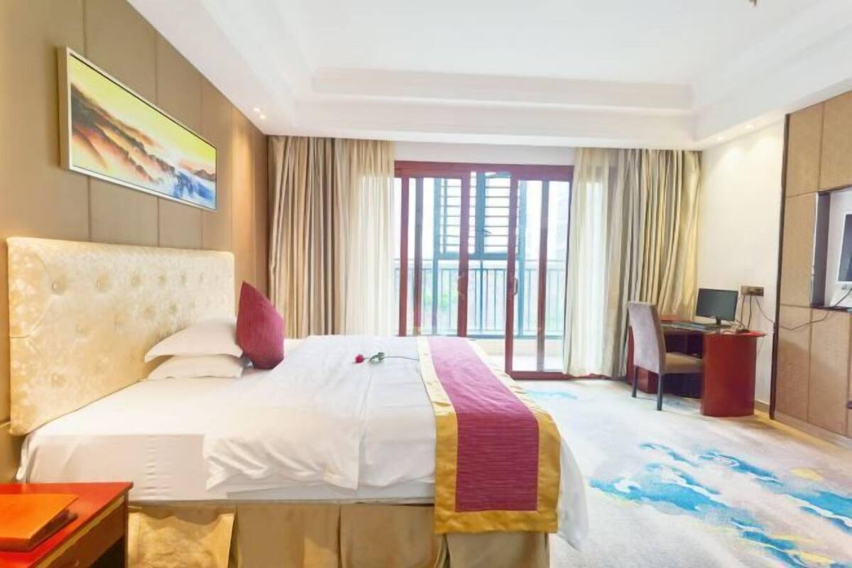 A hotel room with a large bed, a desk with a computer, a TV, and a balcony with curtains.