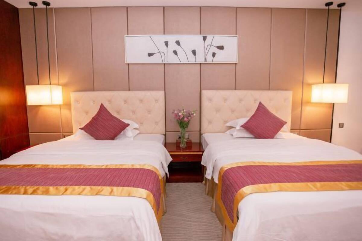 A hotel room with two beds, each with a headboard, a bedside table with a vase, and wall-mounted lamps.
