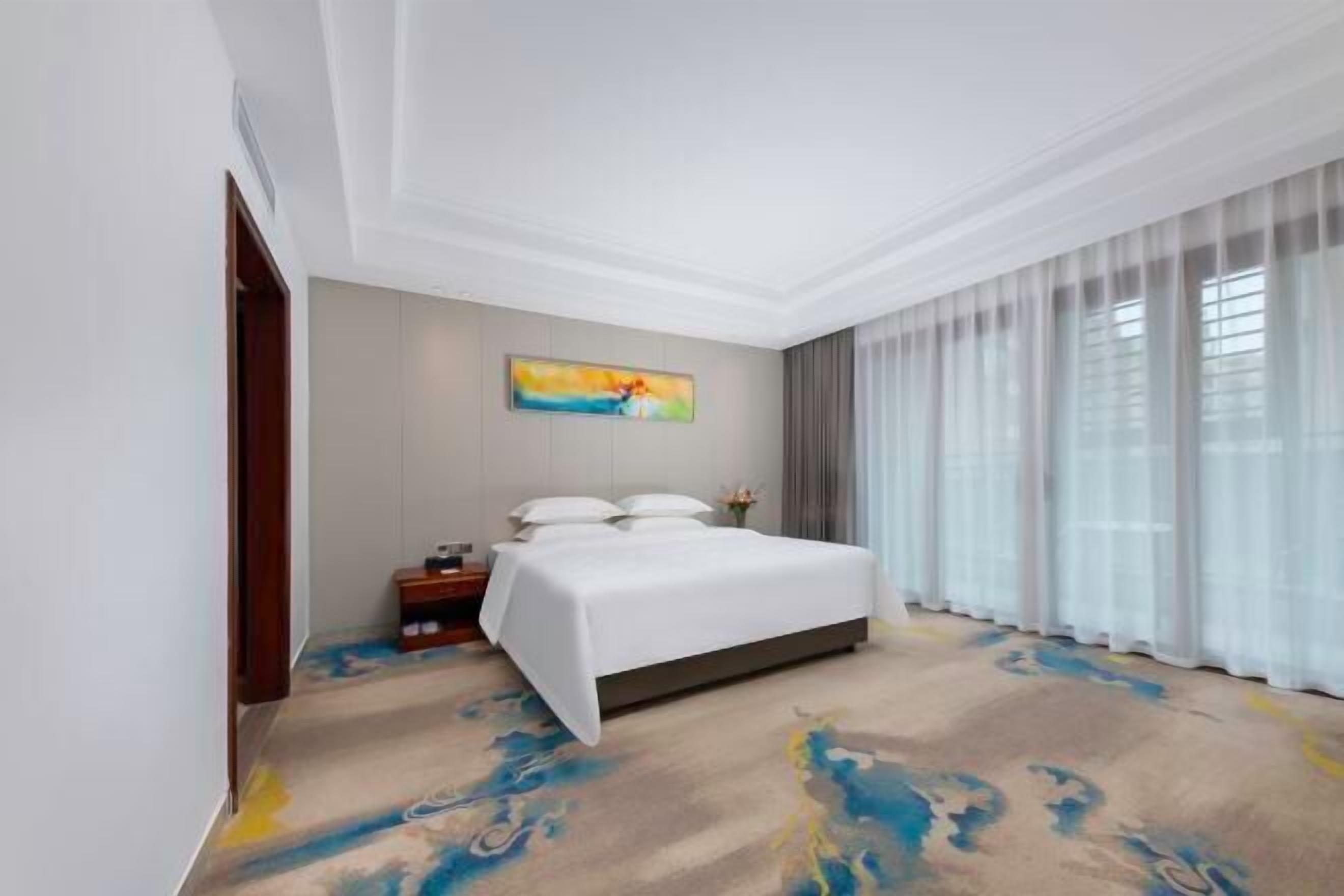 Habitación de hotel con una cama grande, una mesita de noche, un cuadro en la pared y una ventana con cortinas.