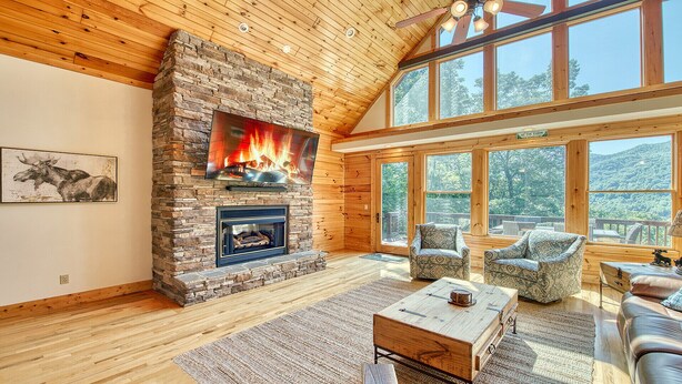 Amplia sala de estar con chimenea de piedra, techo de madera y ventanas grandes que ofrecen vistas al exterior.