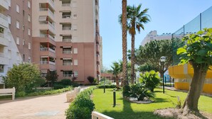 Un immeuble résidentiel doté de nombreux balcons, d’un jardin bien entretenu avec des palmiers et des arbustes, et d’un chemin pavé.