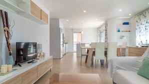 Ein modernes Wohnzimmer mit Flachbildfernseher, Holzsäcken, einem Essbereich mit Stühlen und einer Couch mit gemustertem Kissen.