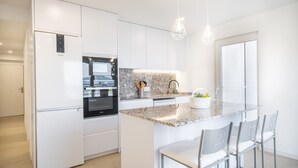 Eine moderne Küche mit weißen Schränken, einem eingebauten Ofen, einem Kühlschrank und einer Kochinsel mit Granitplatte und Barhocker.