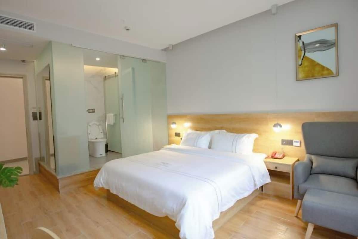 Una habitación de hotel con una cama grande, una mesita de noche con lámpara, una silla y un baño con inodoro y ducha.