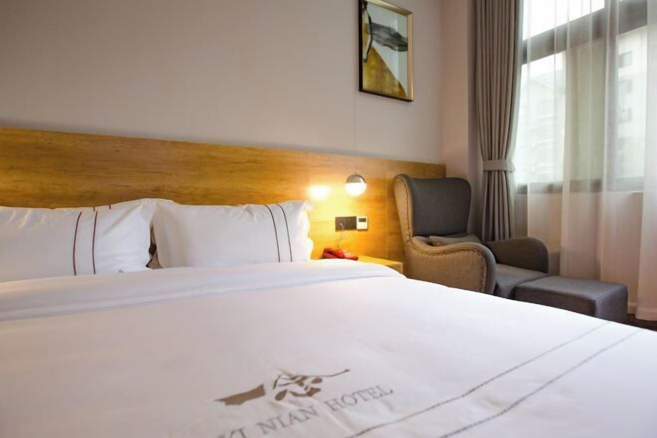 A hotel room with a neatly made bed, a wooden headboard, a wall-mounted lamp, a chair, and a window with curtains.
