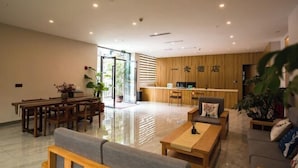 Un hall d’hôtel moderne avec un comptoir d’accueil en bois, un coin salon avec des canapés et une table basse, et un espace repas avec une longue table et des chaises.