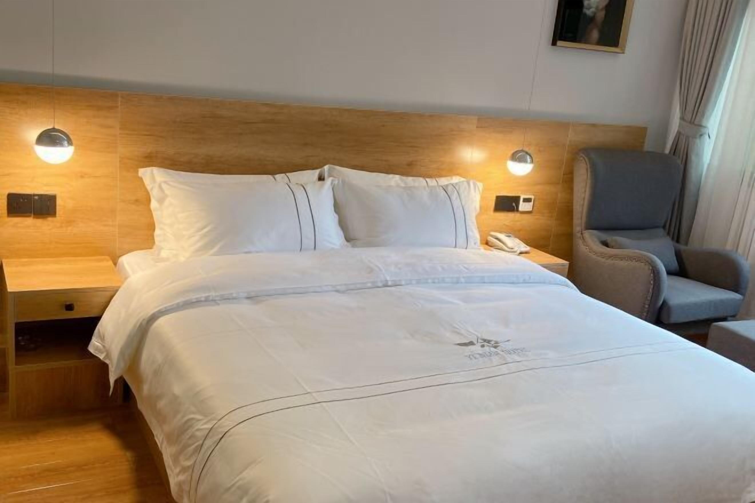 A hotel room with a wooden headboard, a bed with white bedding, a bedside table with a lamp, and a chair.