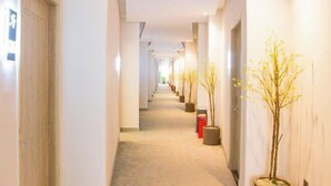 Un couloir d’hôtel avec un sol recouvert de moquette, des murs blancs et des plantes en pot disposées le long des murs.