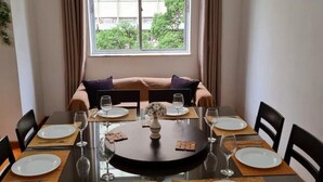 A dining area with a round table set for six, surrounded by chairs. There is a window with curtains, and a sofa is visible in the background.