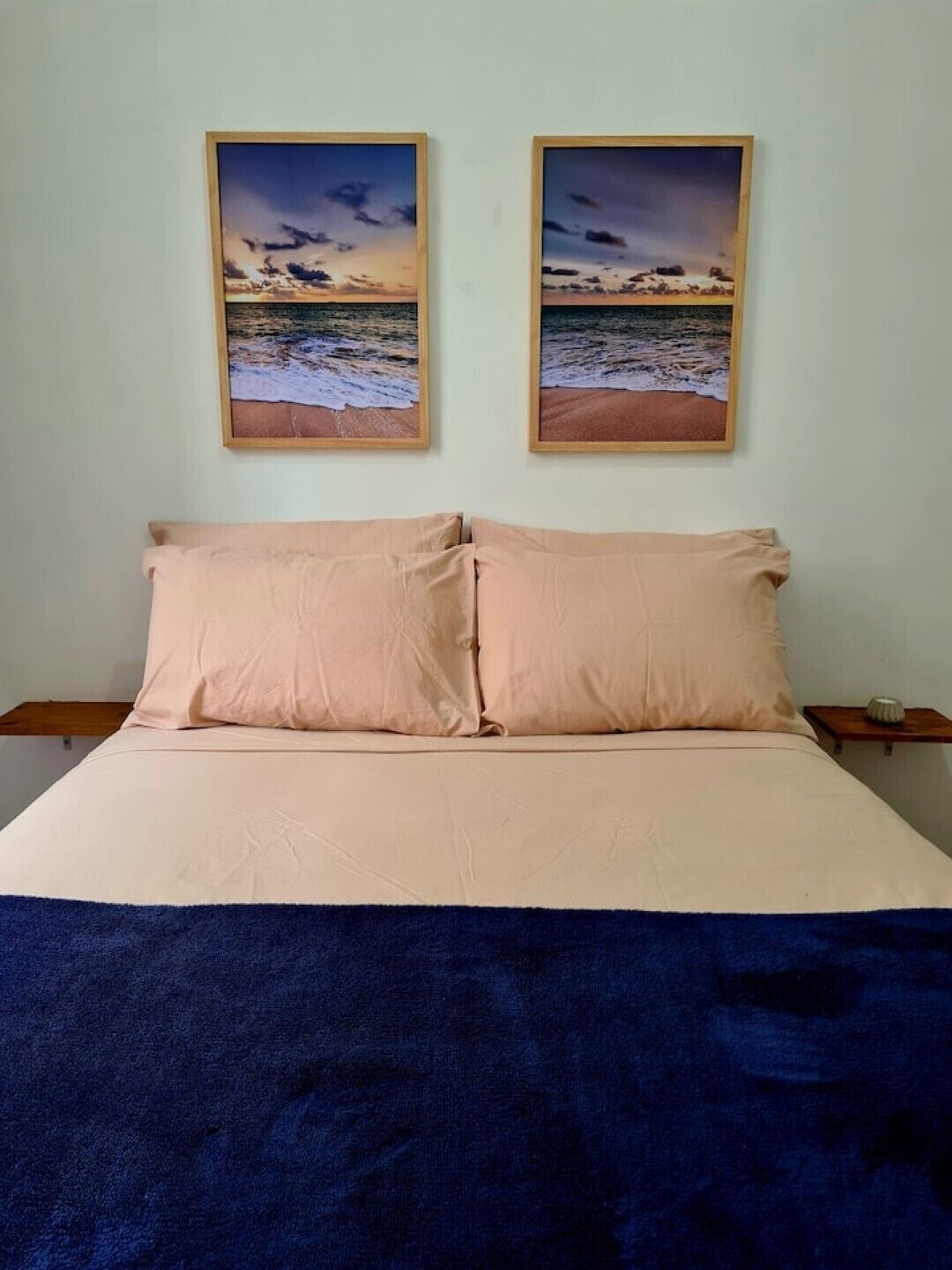 A bed with two pillows, a blue blanket, and two framed pictures of a beach scene on the wall above.