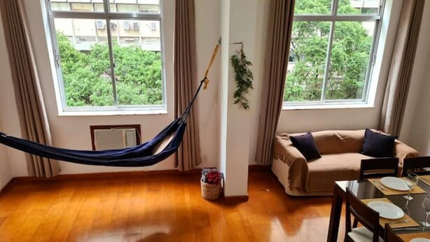 A living room with a hammock, a sofa, a dining table set for two, and large windows with a view of greenery.