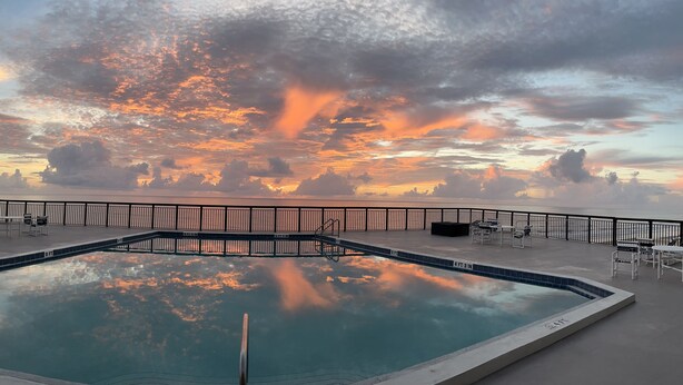 Piscina com vista para o pôr do sol sobre o oceano, rodeada por um deck com cadeiras e guarda-corpo.