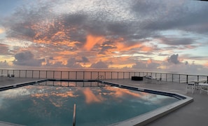 Una piscina con vista al atardecer sobre el océano, rodeada por una terraza con sillas y barandilla.