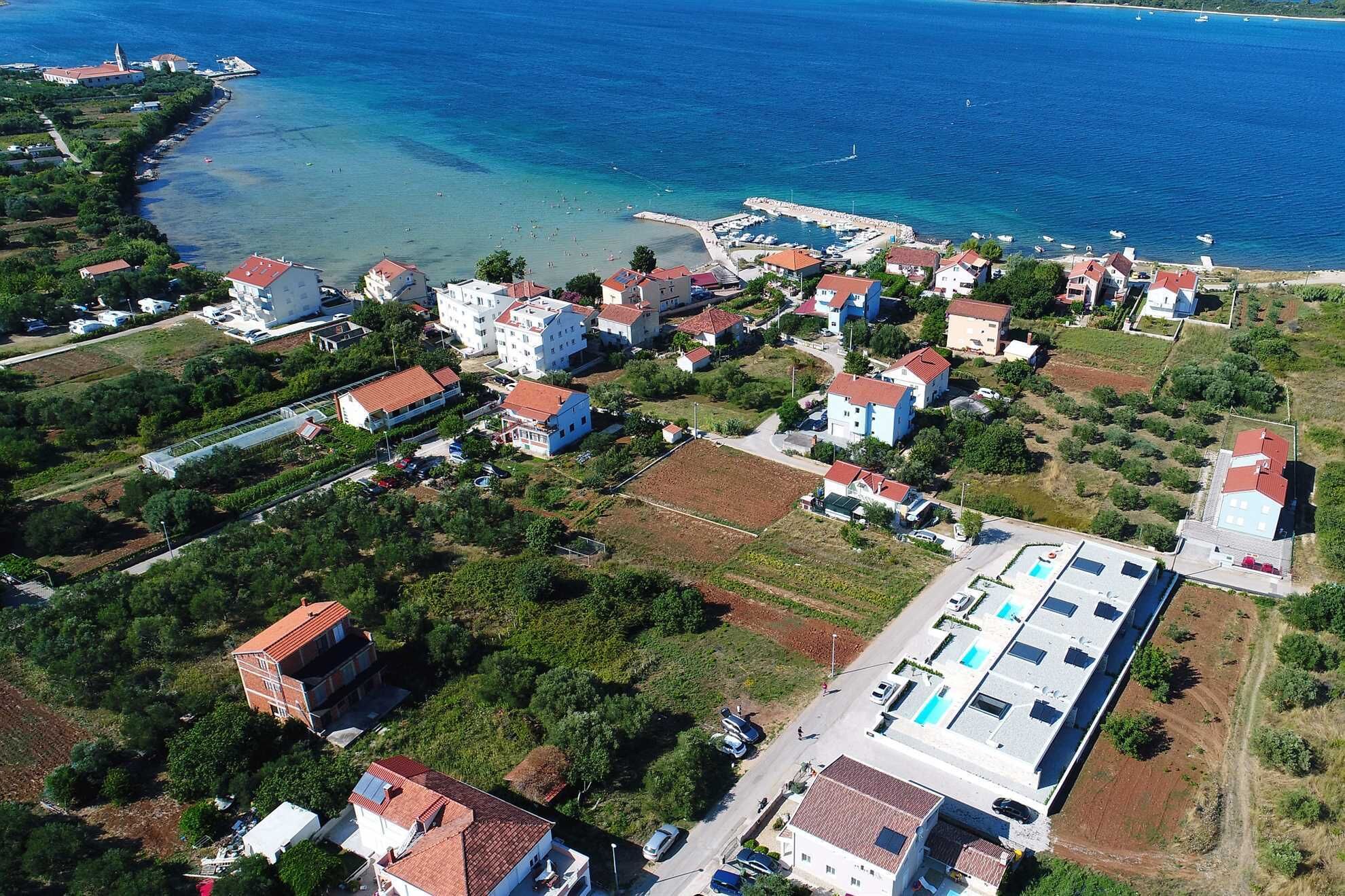 An aerial view of a coastal residential area with houses, a marina, and a large building with a flat roof and multiple windows.