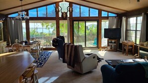 Amplia sala de estar con una gran mesa de comedor, asientos cómodos, televisión y vistas al exterior a través de grandes ventanales.