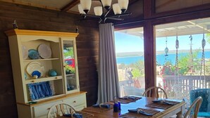 Un comedor con mesa de madera, sillas blancas y una lámpara de araña. Se ve el océano a través de ventanas amplias.
