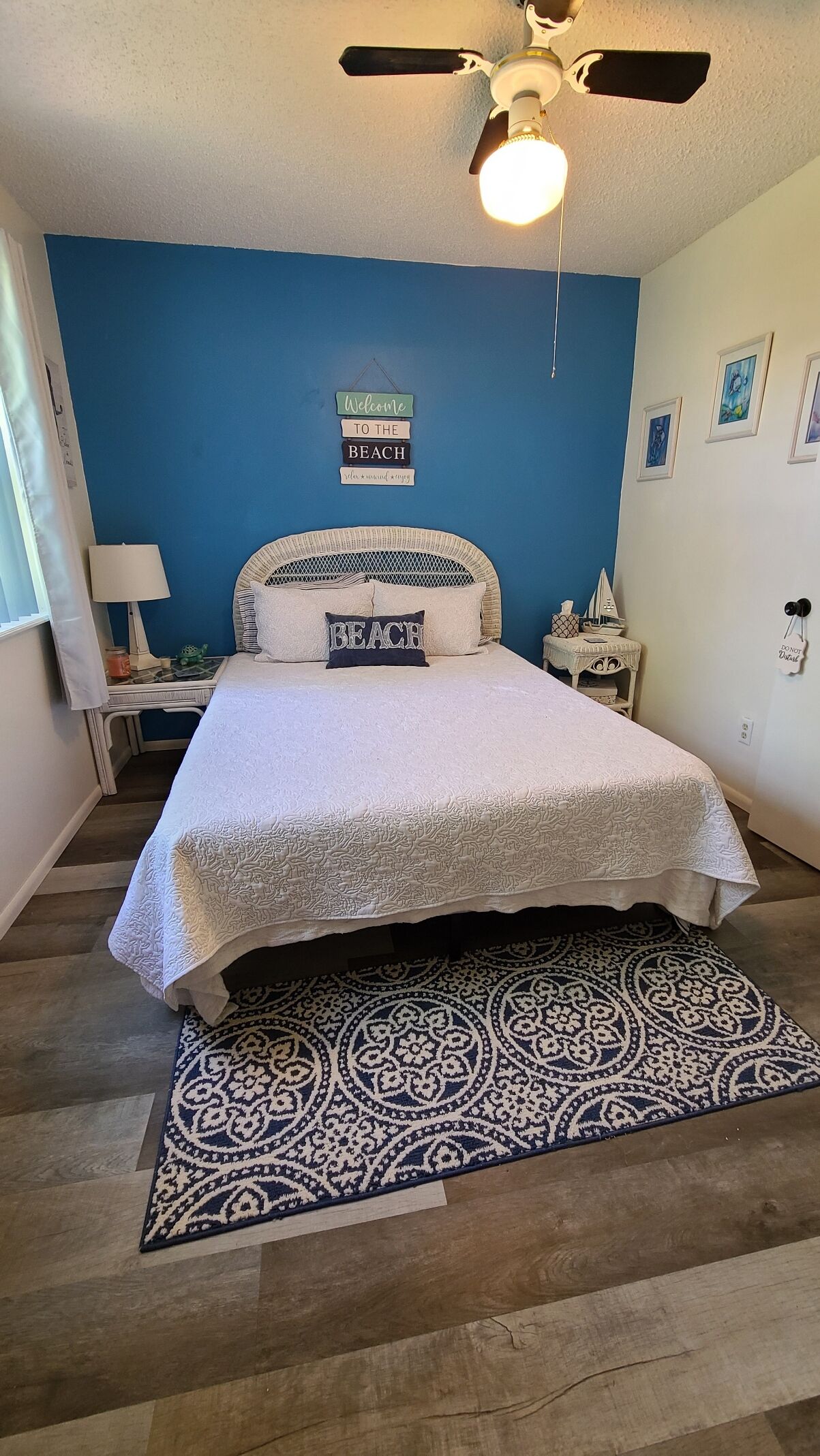 Une chambre à coucher avec un lit, un ventilateur de plafond, un panneau d’accueil, une petite table avec une lampe et un tapis.
