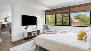Een slaapkamer met een groot raam, een flatscreen televisie, een nachtkastje met een plant en een bed met een decoratief kussen.