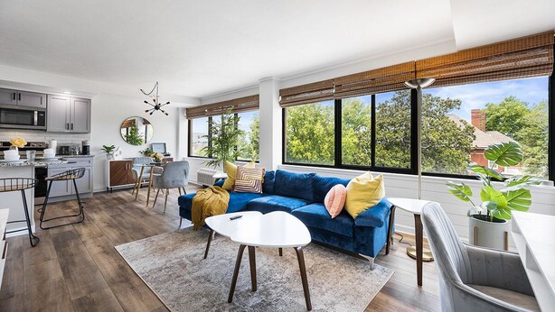 Een moderne woonkamer met een blauwe bank, een witte salontafel en een keukenhoek met grijze kastjes en een magnetron.