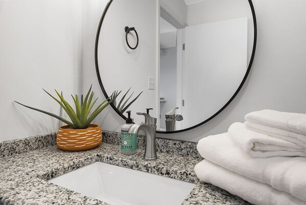Une salle de bain avec un comptoir en granit, un lavabo blanc, un miroir rond, une plante en pot, un distributeur de savon et une pile de serviettes blanches.