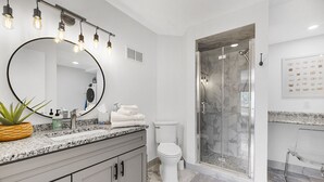 Een moderne badkamer met een grote spiegel, een granieten aanrecht, witte kastjes, een toilet, een douche met glazen wanden en een potplant.