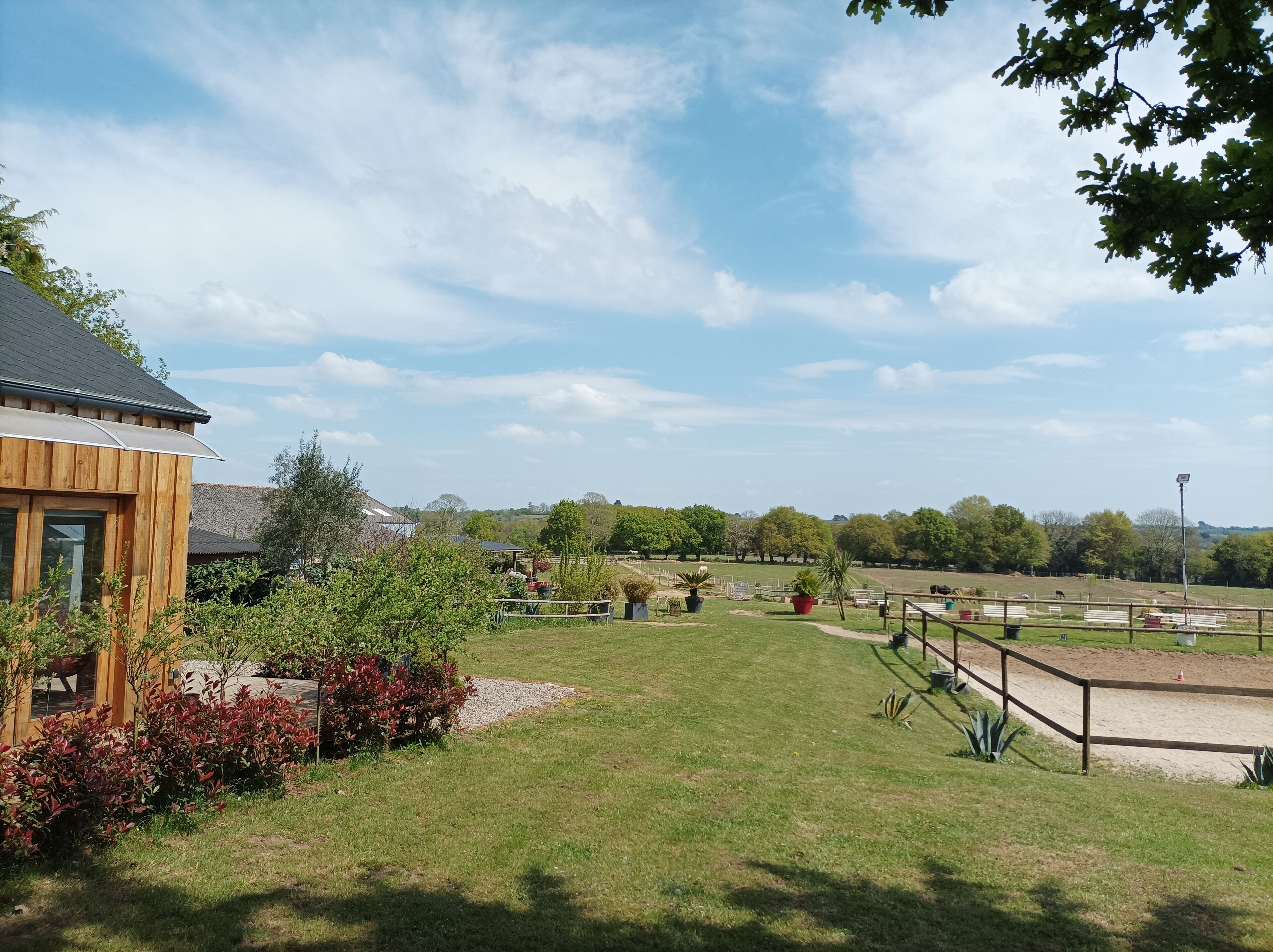 A grassy area with a wooden building, a fenced horse riding area, and trees in the background.
