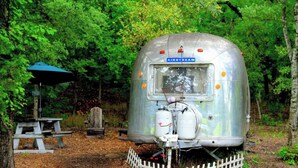 A vintage Airstream trailer with a small window and a metal grill in front, surrounded by a wooded area with a picnic table and a blue umbrella.