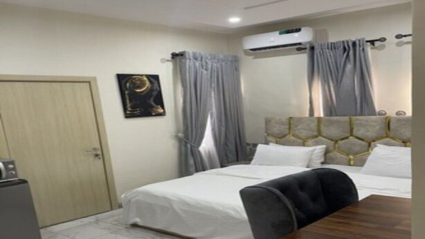 Quarto de hotel com uma cama, uma escrivaninha e uma cadeira. Há um ar condicionado na parede e uma porta à esquerda.