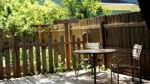A wooden deck with a table and two chairs, surrounded by a wooden fence and greenery.