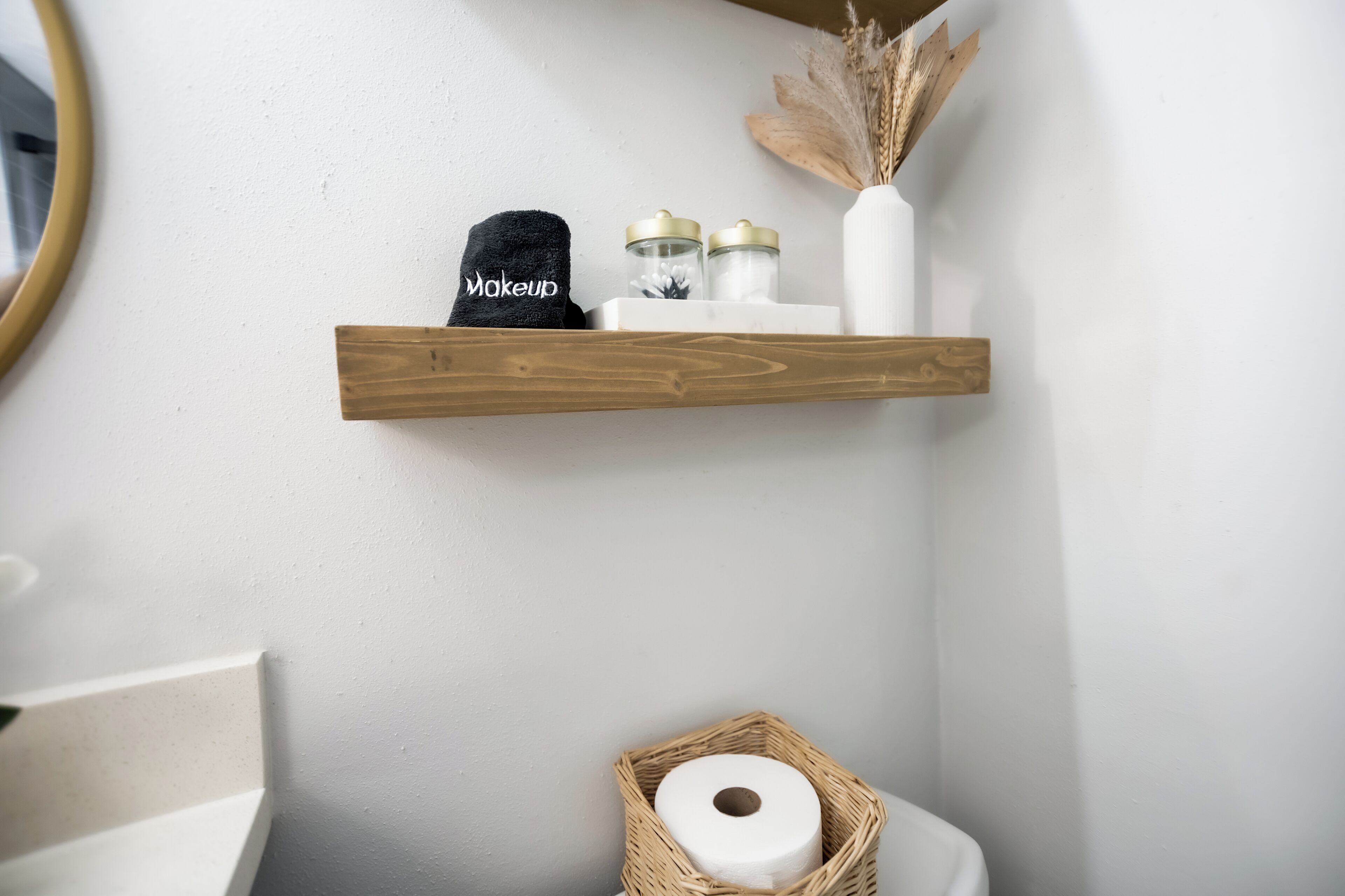 Un baño con un estante de madera que sostiene una toalla negra, un jarrón blanco con plantas secas y dos frascos con tapas doradas.