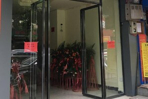 Entrada de hotel con puerta de cristal, letrero rojo y plantas en macetas.