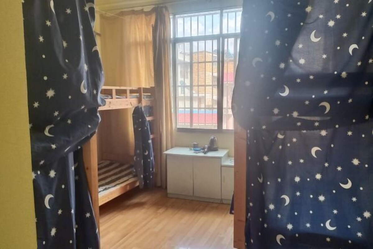 Quarto com beliche, uma pequena escrivaninha e cadeira, e uma janela com grades.