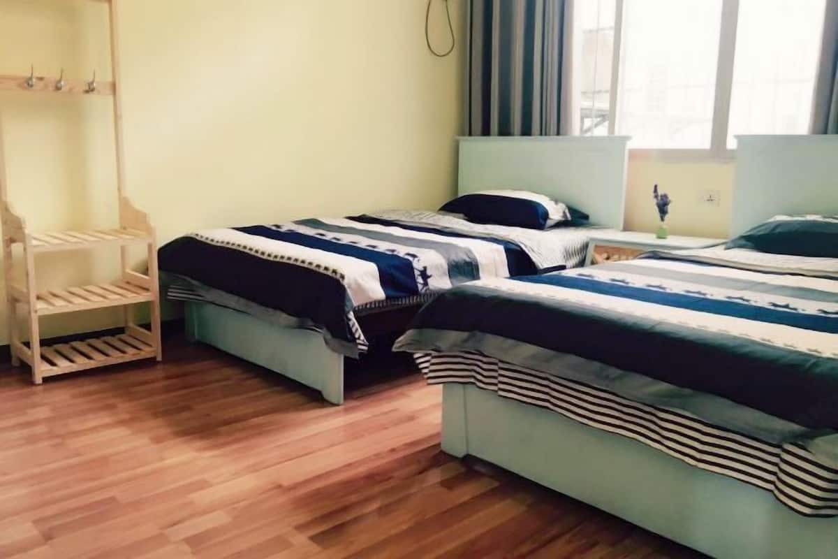 Una habitación con dos camas, una estantería de madera y un ventanal con cortinas.