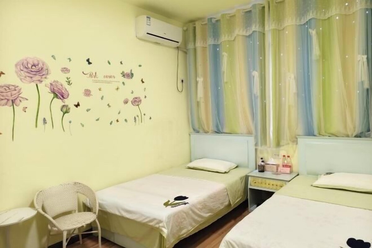 Quarto com duas camas de solteiro, uma cadeira, uma mesinha de apoio com produtos de higiene e uma janela com cortinas finas.