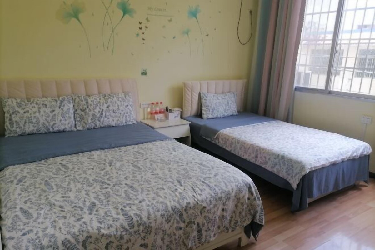 Quarto com duas camas, cada uma com roupa de cama e almofadas estampadas, uma pequena mesa de cabeceira com abajur e uma janela com cortinas.