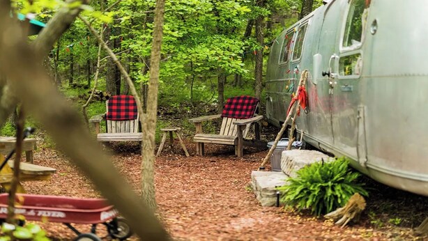 A rustic campsite with a vintage camper parked among trees, two Adirondack chairs with plaid covers, and a small table.