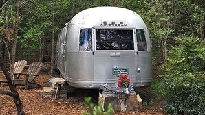 A vintage Airstream trailer parked in a wooded area with a bench nearby.