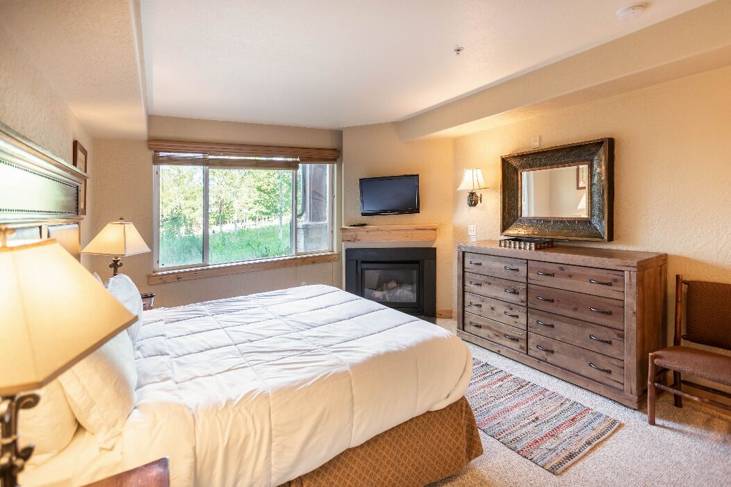 A bedroom with a bed, a dresser, a television, a fireplace, and a window with a view of greenery.