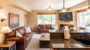 Una sala de estar con un sofá de cuero marrón, una mesa de centro y un televisor montado en la pared. Hay una zona de cocina con encimera de granito y lavavajillas.