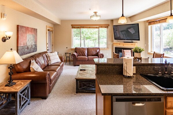 A living room with a brown leather sofa, a coffee table, and a TV mounted on the wall. There is a kitchen area with a granite countertop and a dishwasher. - Quiet condo with free parking, pool, hot tub & sauna. 7 min walk to gondola.  (Park City)