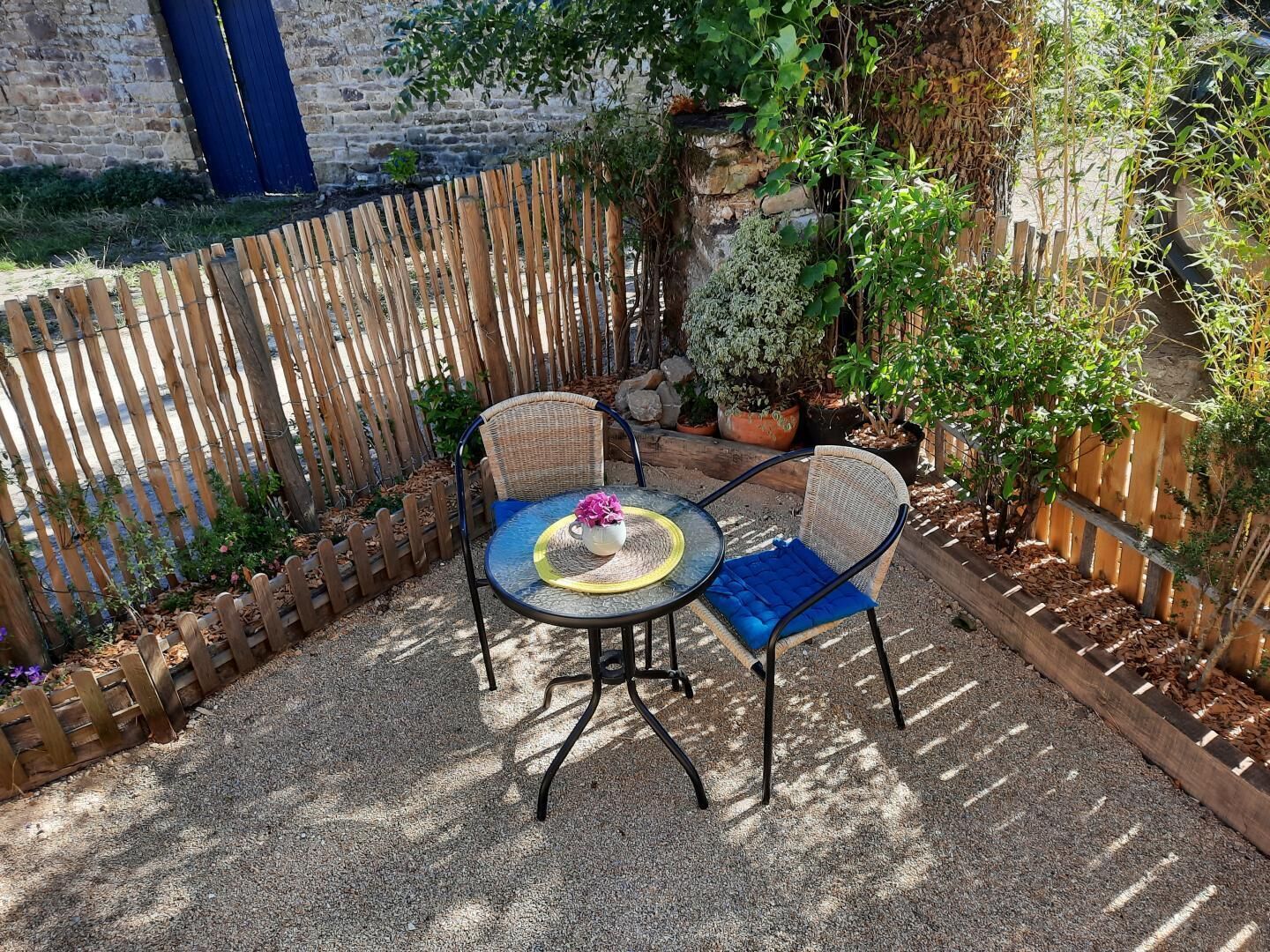 Une terrasse extérieure avec deux fauteuils en osier et une table ronde, entourée d’une clôture en bois et de différentes plantes en pot.