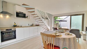 A modern kitchen with white cabinets, a built-in oven, and a microwave. There is a dining area with a round table, chairs, and a staircase leading to an upper level. - Steredeg - BADEN - Gîte (BADEN)